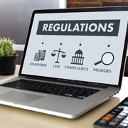 A laptop on a table with the word “regulations” on the screen along with images representing standards, laws, compliance and policies.